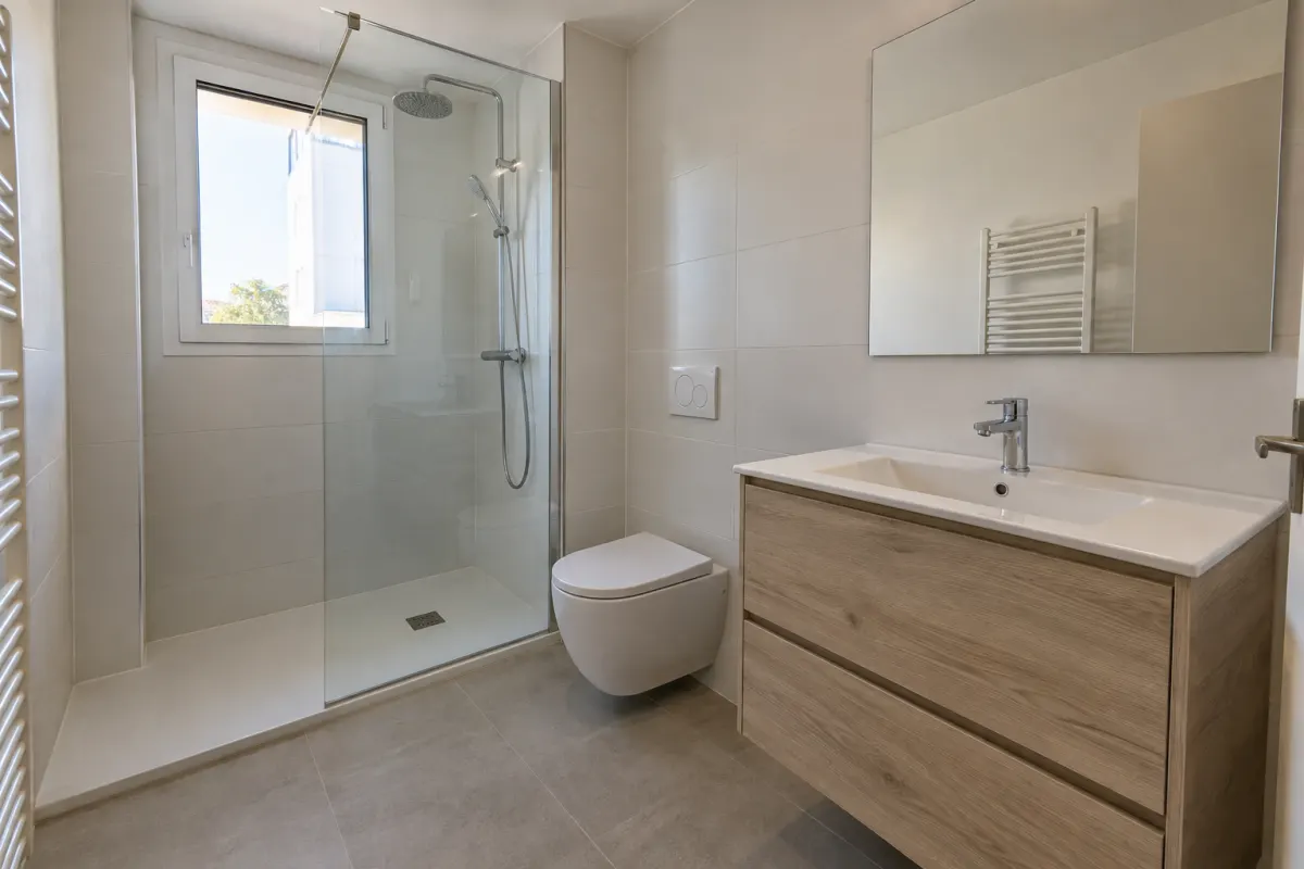 Baño reformado con ducha y mampara de cristal, inodoro suspendido y mueble de lavabo de madera clara (imagen generada con IA)